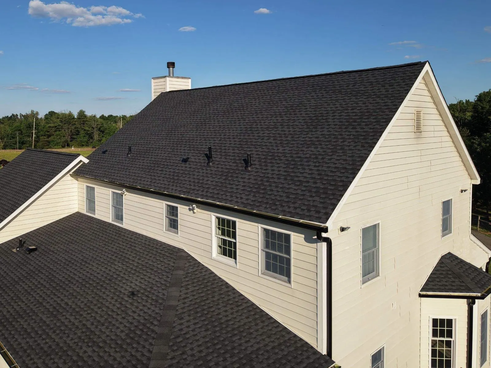 A new GAF Timberline Ultra HDZ roof in Charcoal and black seamless gutters on a cream farmhouse in Skillman, Montgomery Township, seen from above with open fields behind, by Pujols Roofing.