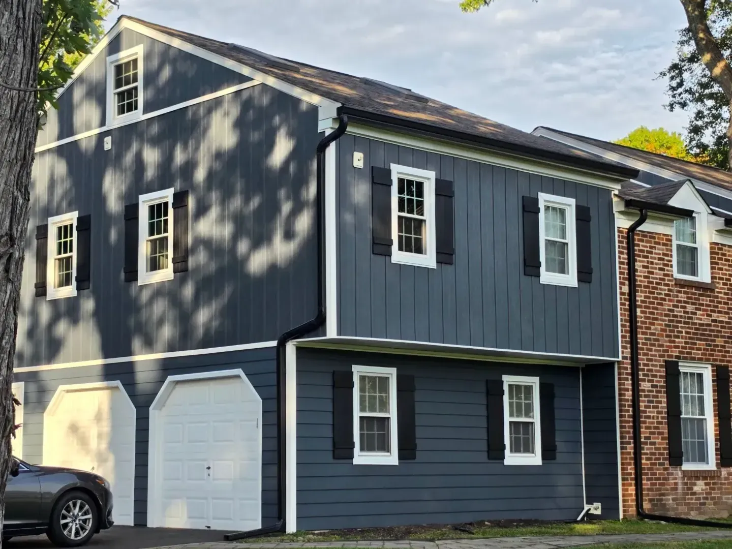 The garage side of a Richboro, PA home with new Everlast composite siding in Blue Spruce, white-trimmed windows with black shutters, black gutters and downspouts, and a brick section, by Pujols Roofing.