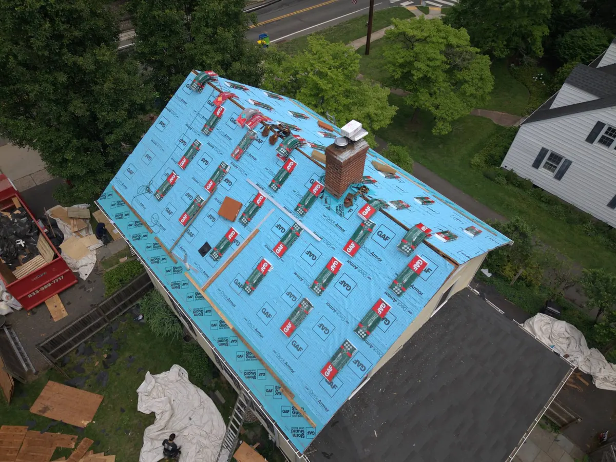 Drone view of a Rocky Hill, NJ roof mid-replacement: new plywood under blue GAF Deck-Armor underlayment, bundles of shingles staged, the old brick chimney in place, by Pujols Roofing.