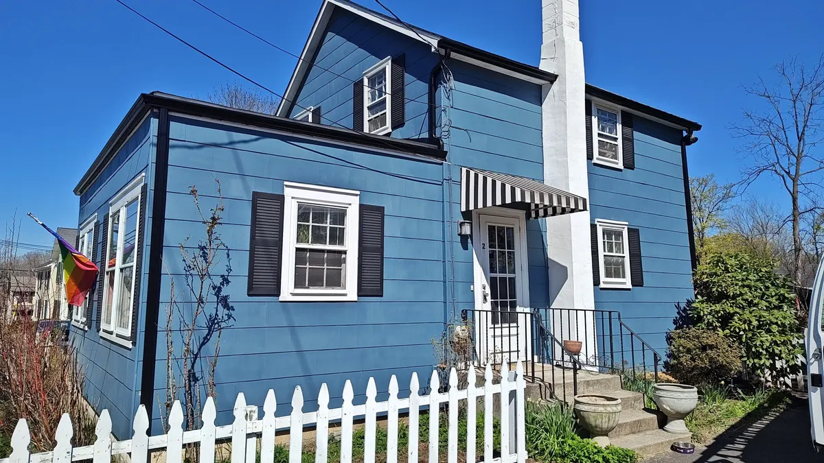 A blue two-story house on Princeton Avenue in Rocky Hill, NJ with a new roof and gutters behind a white picket fence, by Pujols Roofing.